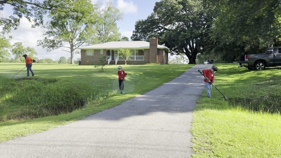 All three brothers trimming the driveway edges in unison, face shields on, at a brick ranch home